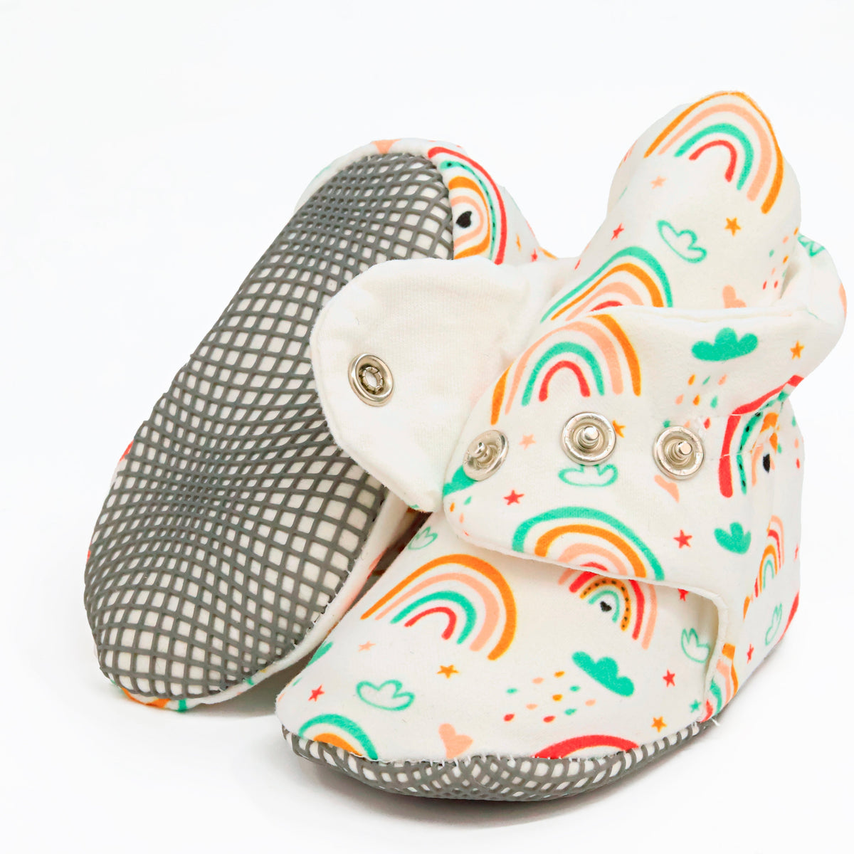 Cream baby booties with colorful rainbows and snap straps, showing one sole and an open snap strap on white.