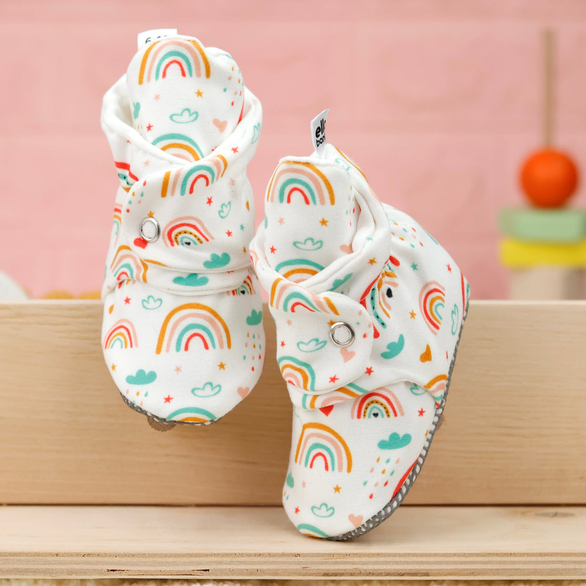 Front view of a pair of cream baby booties with colorful rainbows and snap straps.