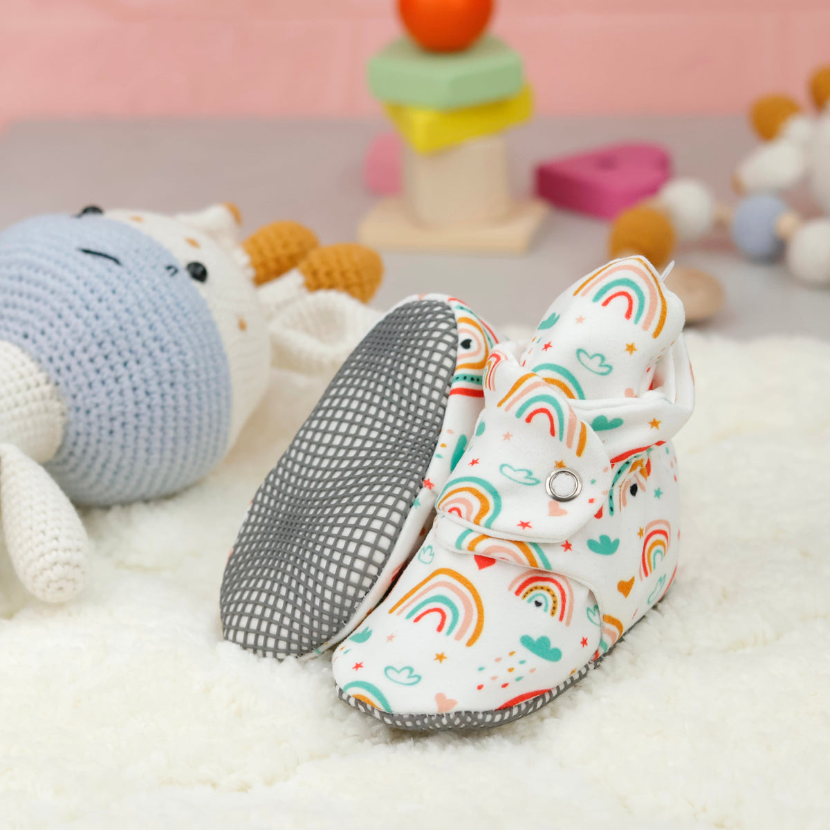 Cream baby booties with colorful rainbows and snap straps arranged on a surface with one sole facing the camera.