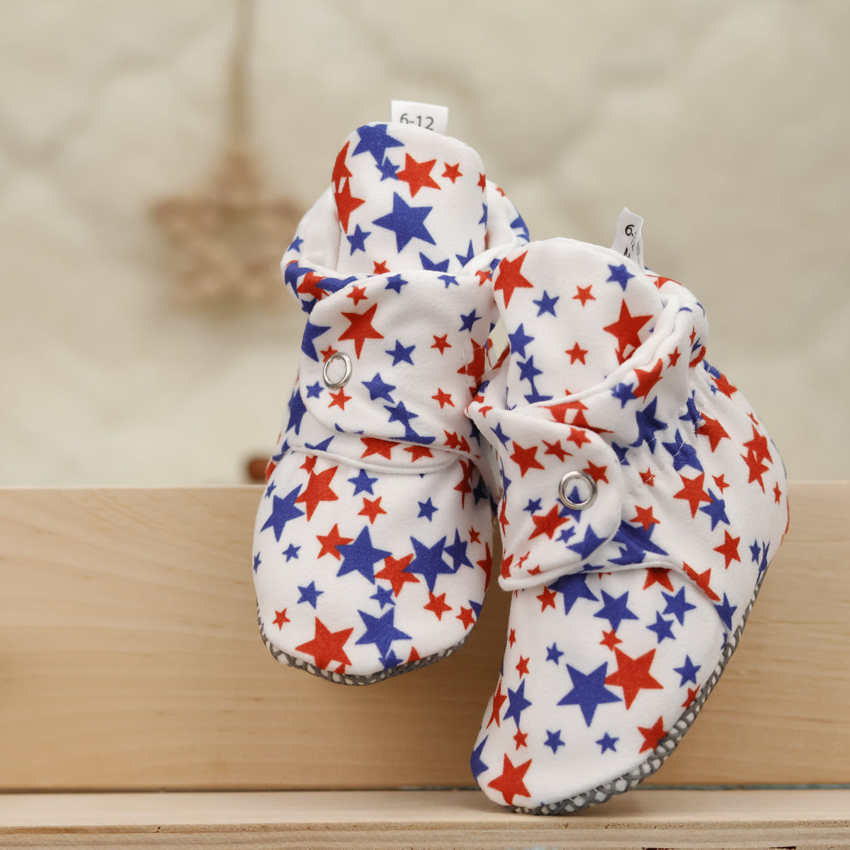Front view of a pair of white baby booties with red-and-blue stars and snap straps.