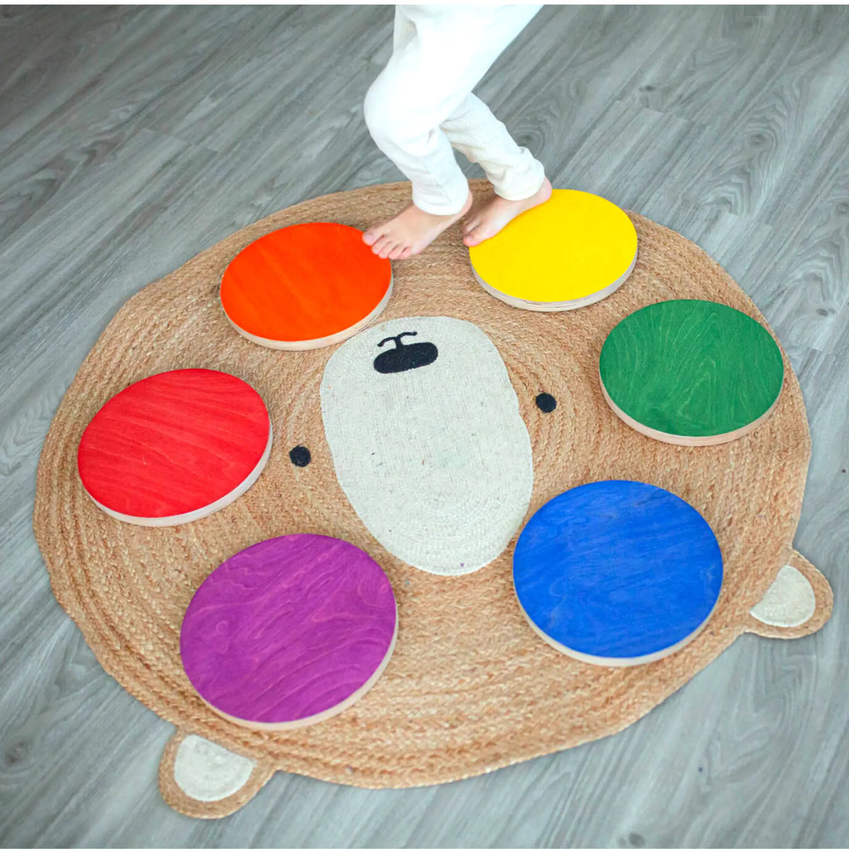 Child stepping among six rainbow-colored round stepping stones arranged on a bear-shaped rug.