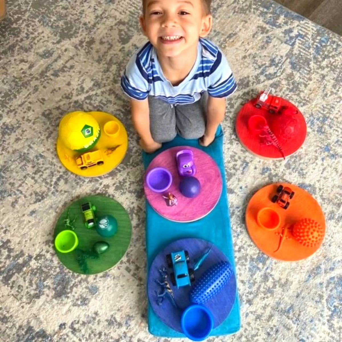 Child seated among rainbow stepping stones topped with color-matched toy cars, cups and other toys.