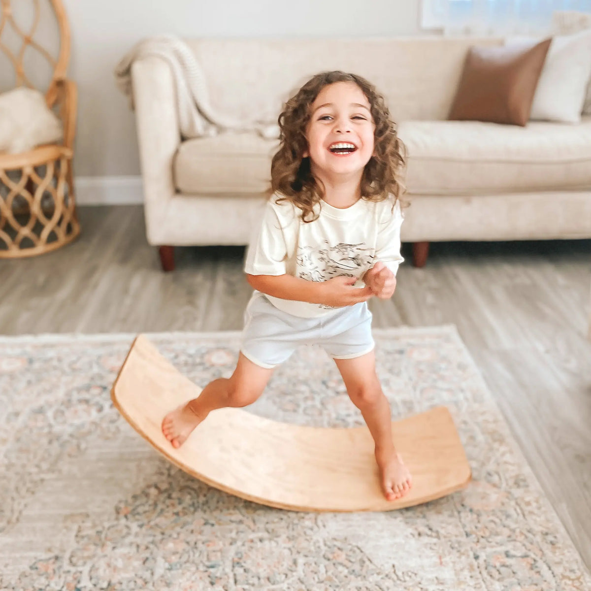 Child standing barefoot on the concave side of a natural-tone wobble board in a living room.