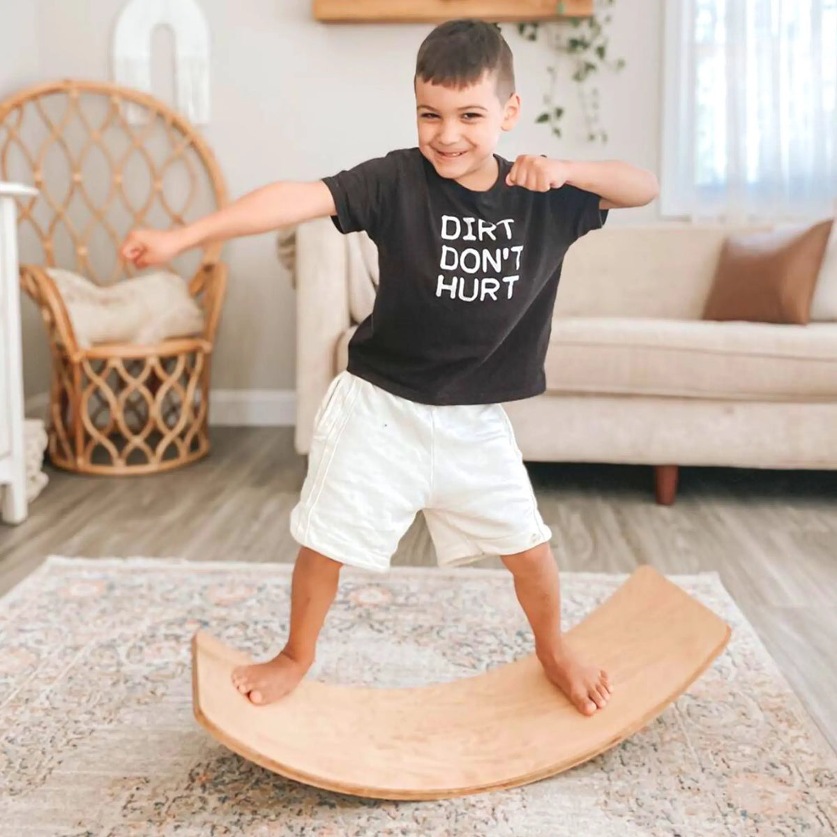 Child standing with arms extended on the concave side of a natural-tone wobble board.